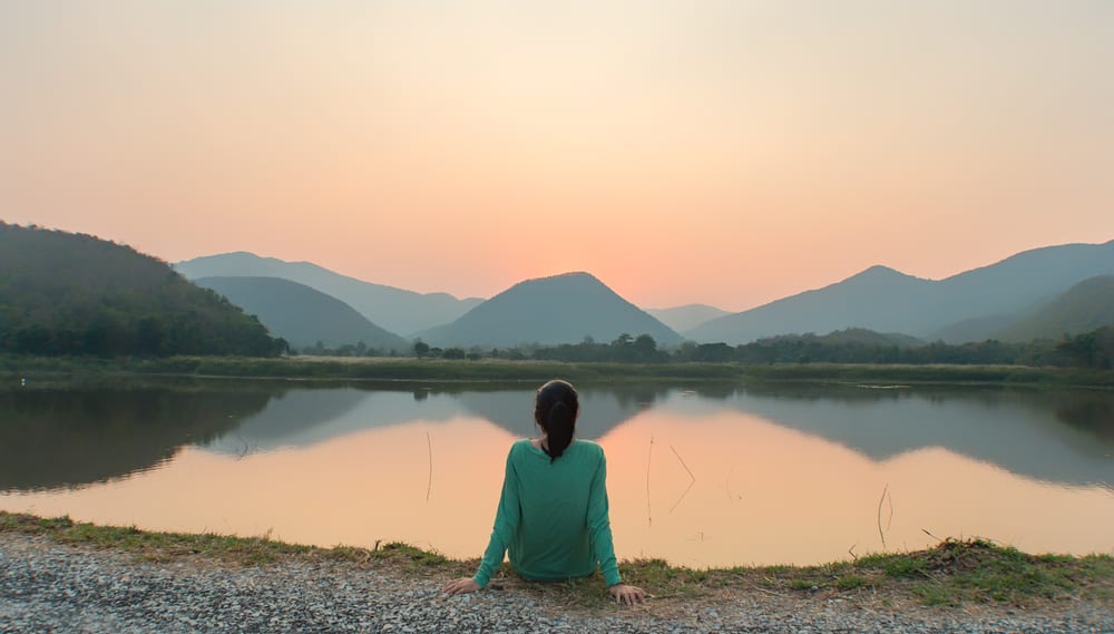 girl sits in front of beautiful lake with mountains in front also sunrise or sunset reflection of sky blue sweater